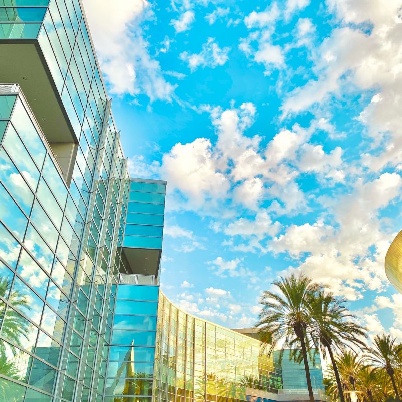 The image shows a modern glass building, palm trees, and a blue sky with scattered clouds in a vibrant, sunny setting.