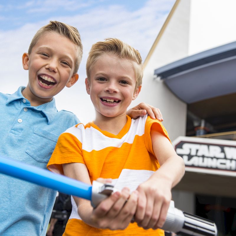 Two smiling boys are standing outside, one holding a blue lightsaber toy. The sign behind them reads "Star Wars Launch Bay."