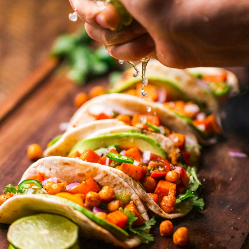 The image shows a hand squeezing lime juice over a row of tacos filled with vegetables and chickpeas, garnished with cilantro on a wooden board.