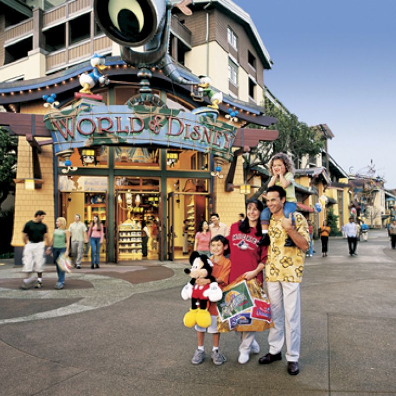 A family poses with a Mickey Mouse toy in front of a store named "World of Disney" at a Disney park, surrounded by other visitors and buildings.