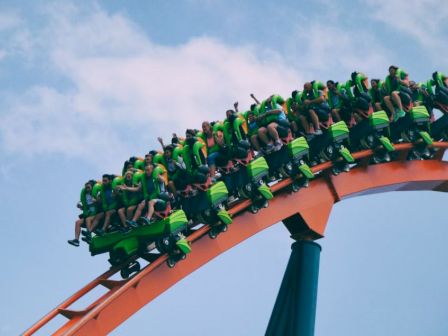 People ride a green roller coaster, ascending a track with blue sky and clouds in the background.