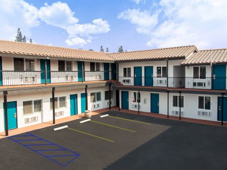 The image shows a two-story motel with blue doors and windows, surrounding a parking lot with spaces and a handicap spot, under a blue sky.