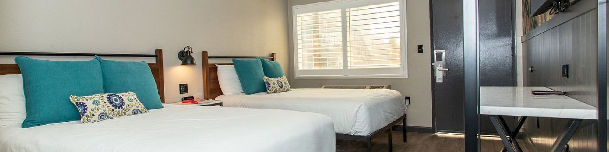 This image shows a modern hotel room with two double beds, teal and floral pillows, a desk, a wall-mounted TV, and a window with blinds.