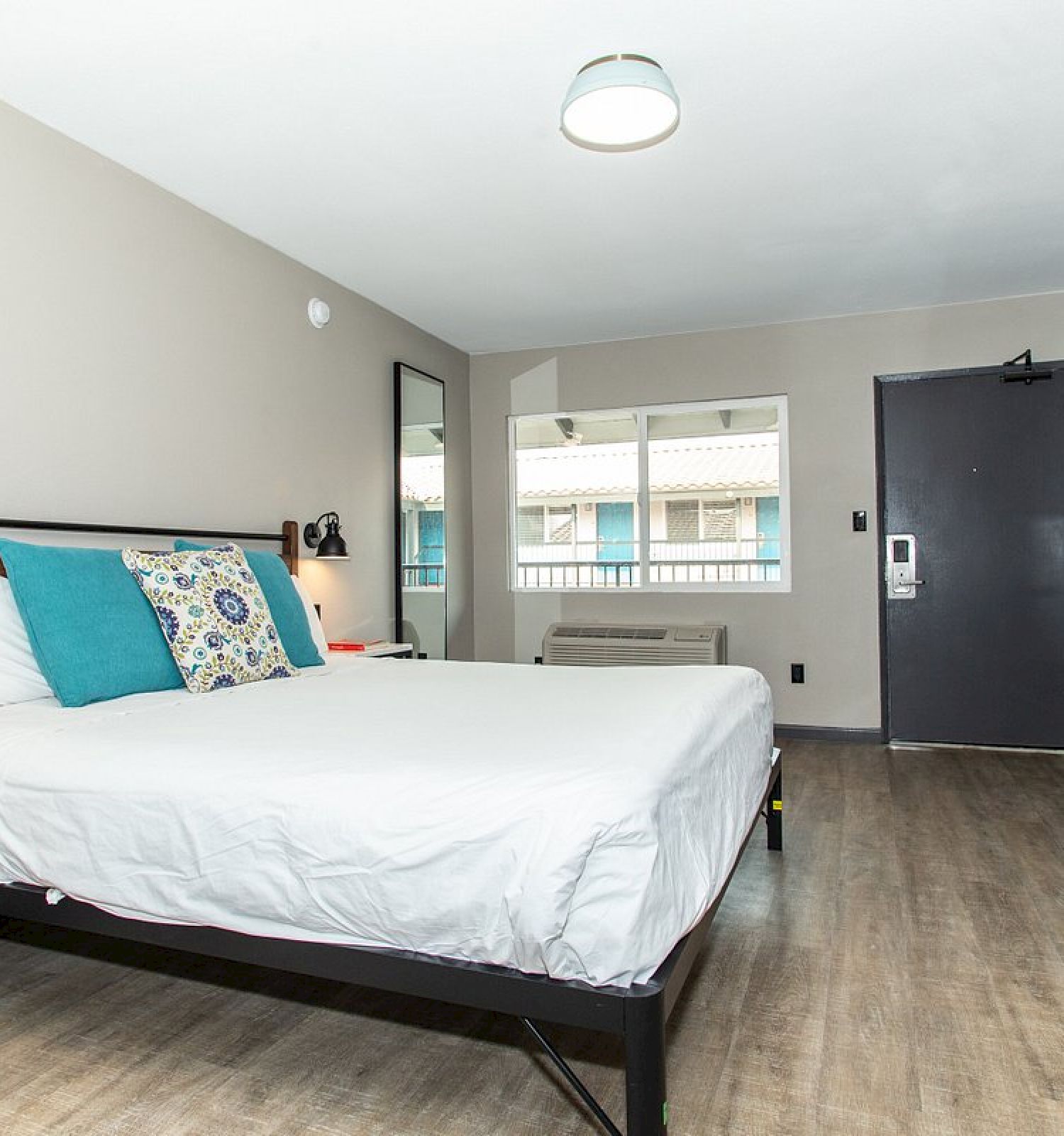 A clean, modern hotel room with a bed, pillows, a small desk, and a window. The room features wooden flooring and a door opposite the bed.