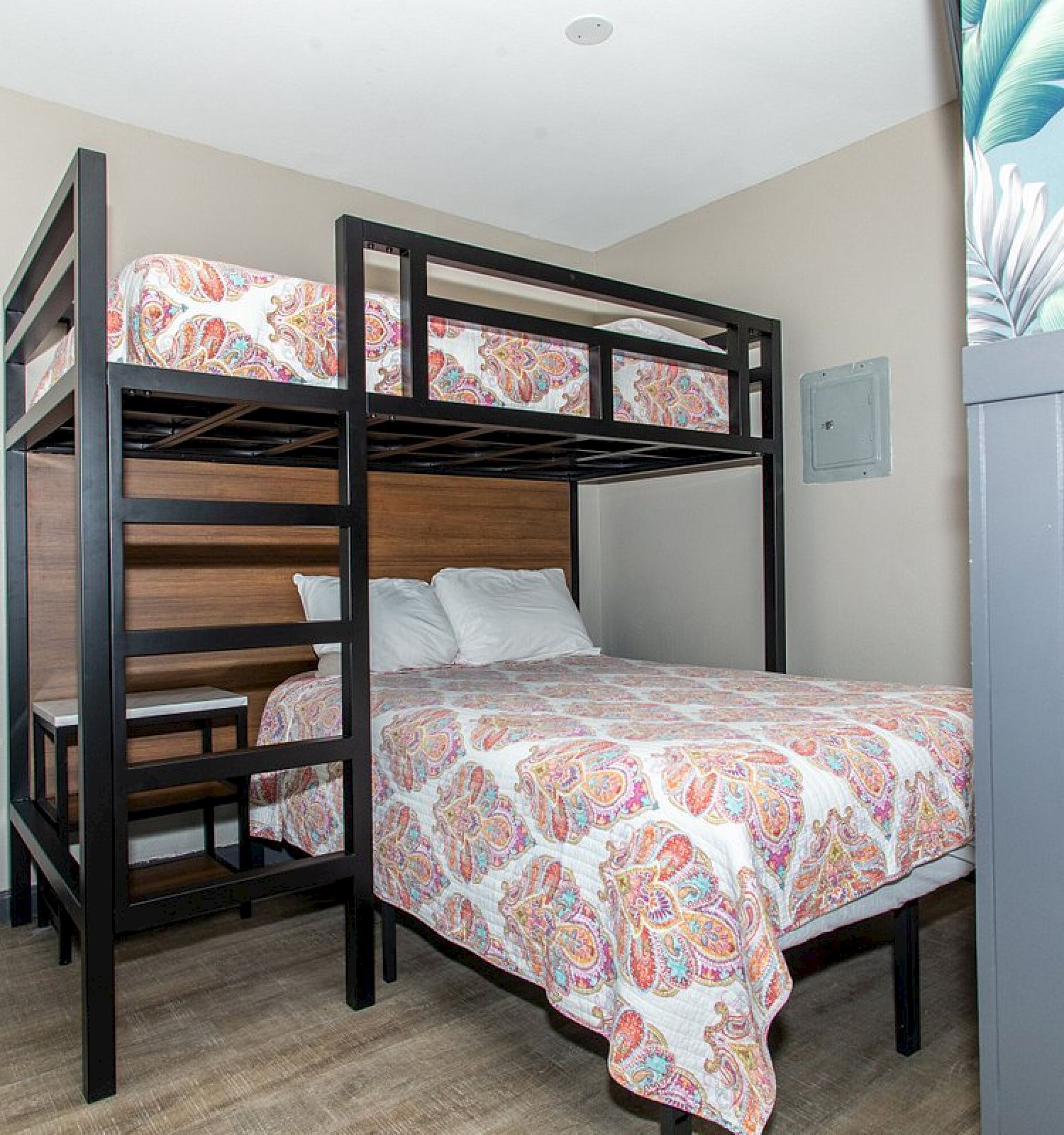 This image shows a bedroom with a bunk bed; the lower bed is larger than the upper bed, both covered with colorful patterned bedspreads.