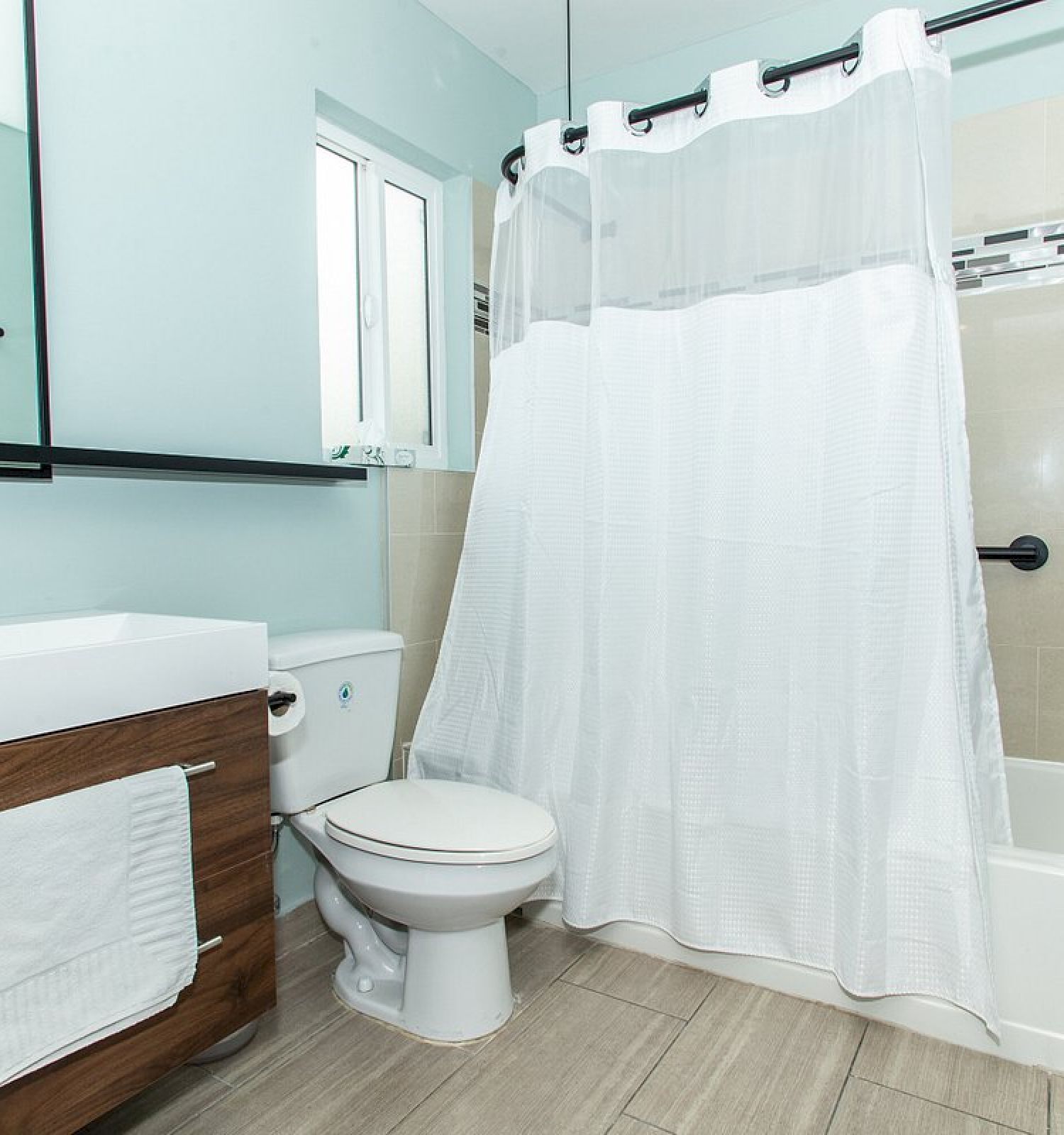 A modern bathroom features a sink with a towel rack, a toilet, a bathtub with a white shower curtain, and a large mirror on the wall.