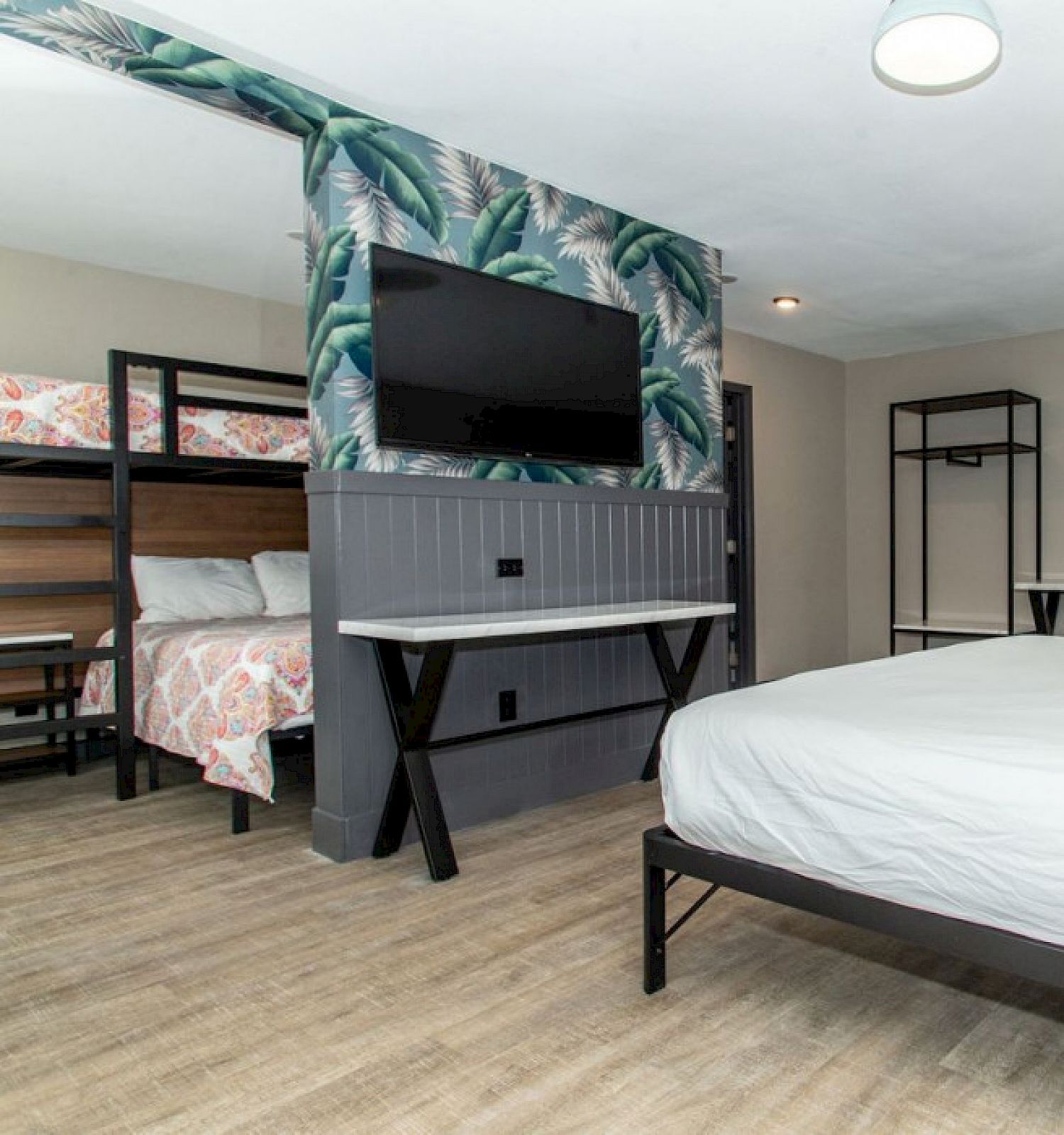 A hotel room with a queen bed and bunk beds. There's a TV mounted on the wall with tropical wallpaper, and two desks at the back of the room.