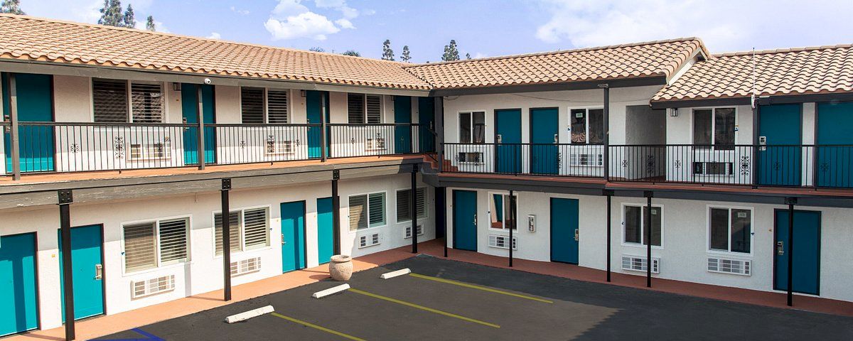 A white building with blue doors and a brown roof, featuring a balcony and a parking lot.