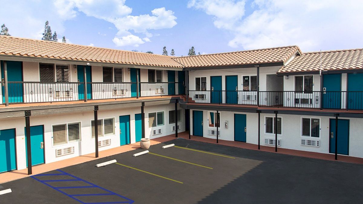The image shows a two-story motel with blue doors, an empty parking area, and a blue sky background ending the sentence.