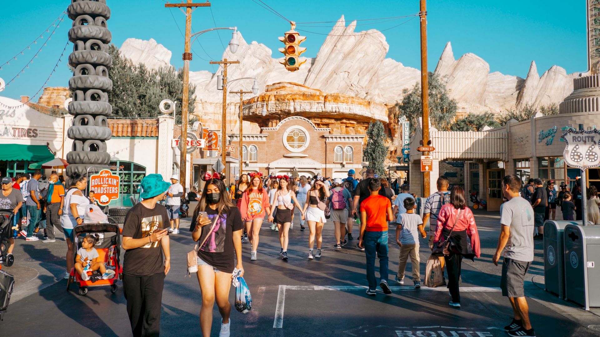 People are walking in an amusement park with themed buildings and attractions under a clear blue sky. The scene is bustling with activity.