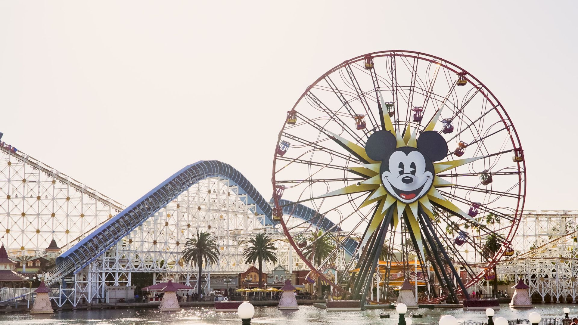 The image shows a theme park with a large Ferris wheel featuring a Mickey Mouse face and a roller coaster in the background, surrounded by water.