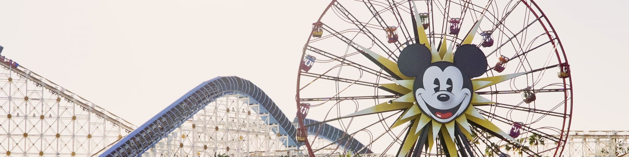 The image shows a theme park with a large Ferris wheel featuring a Mickey Mouse face and a roller coaster in the background, surrounded by water.