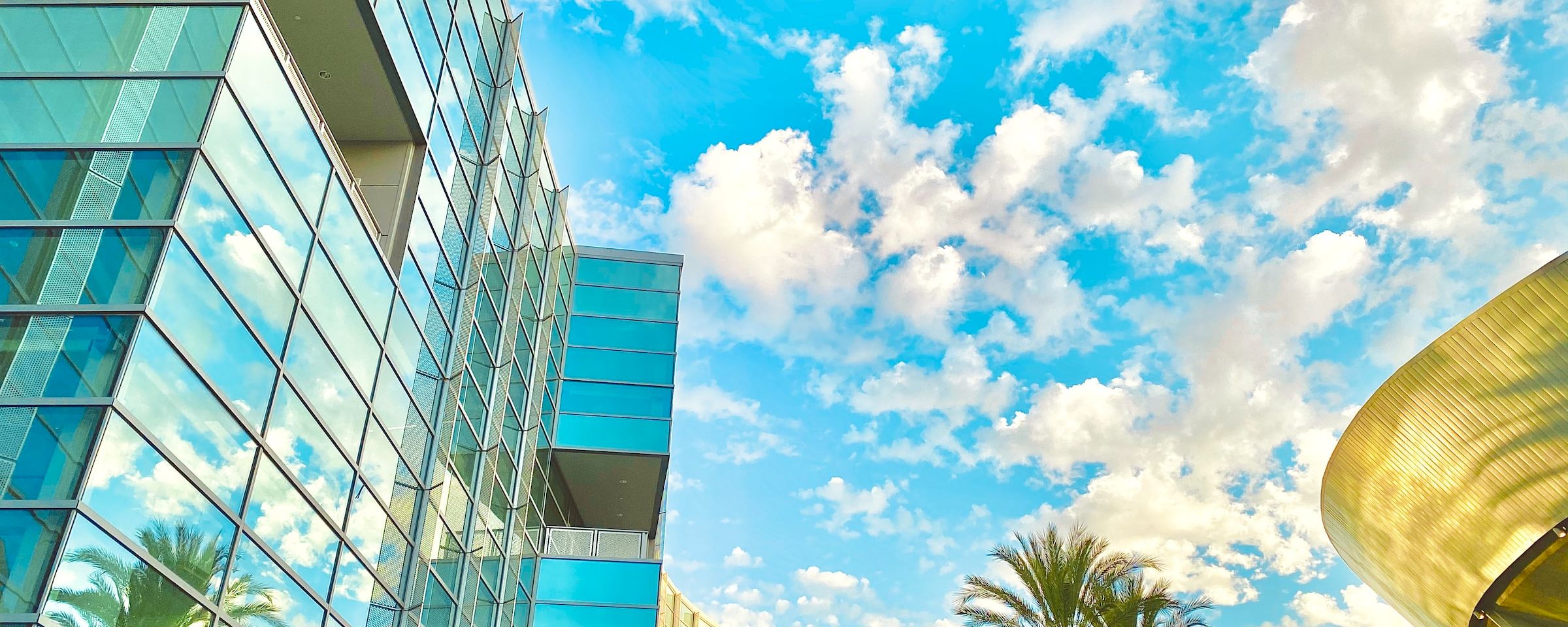A low-angle shot of a building's exterior with a glass facade and a curved roof, set against a blue sky with clouds and palm trees.