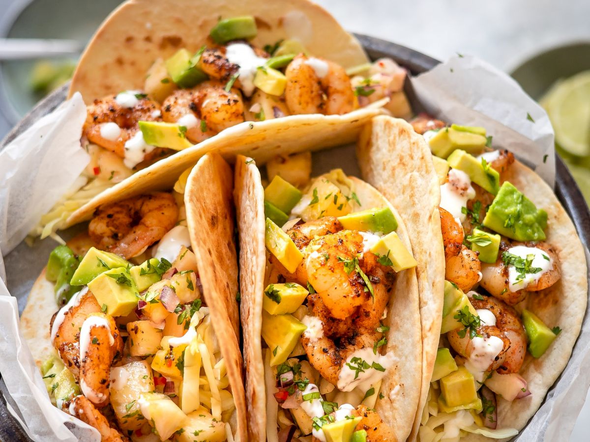 A hand holding a plate with three shrimp tacos topped with diced avocado, cilantro, and a drizzle of white sauce.