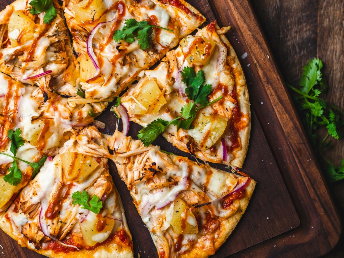 A delicious pizza topped with cheese, chicken, pineapple, red onion, and cilantro sits on a wooden cutting board, with fresh cilantro nearby.