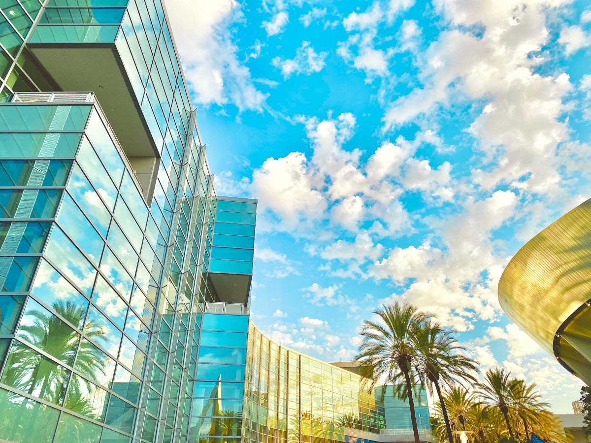 Modern glass buildings with reflections of palm trees, a bright blue sky with scattered clouds, and a golden cylindrical structure on the right.