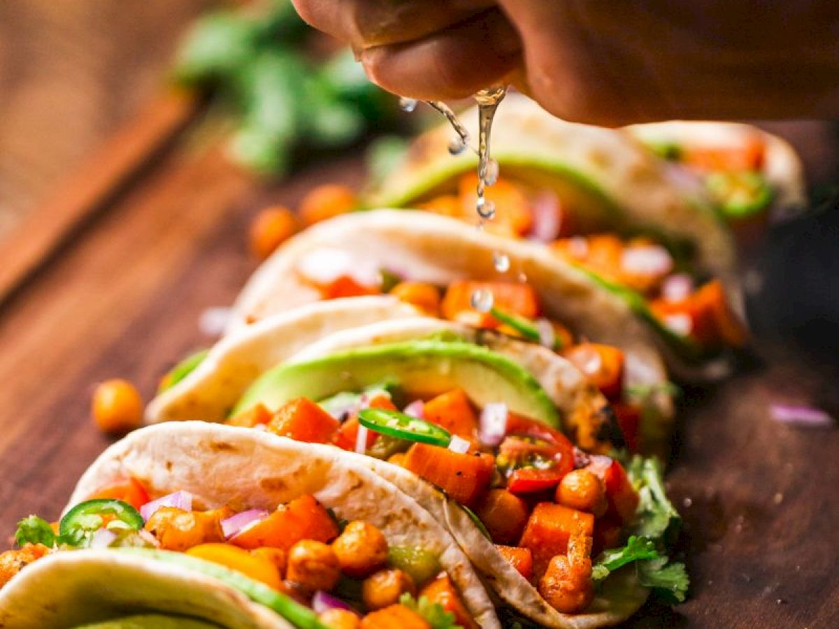 The image shows a hand squeezing lime juice over several tacos filled with chickpeas, diced vegetables, cilantro, and avocado slices on a wooden board.