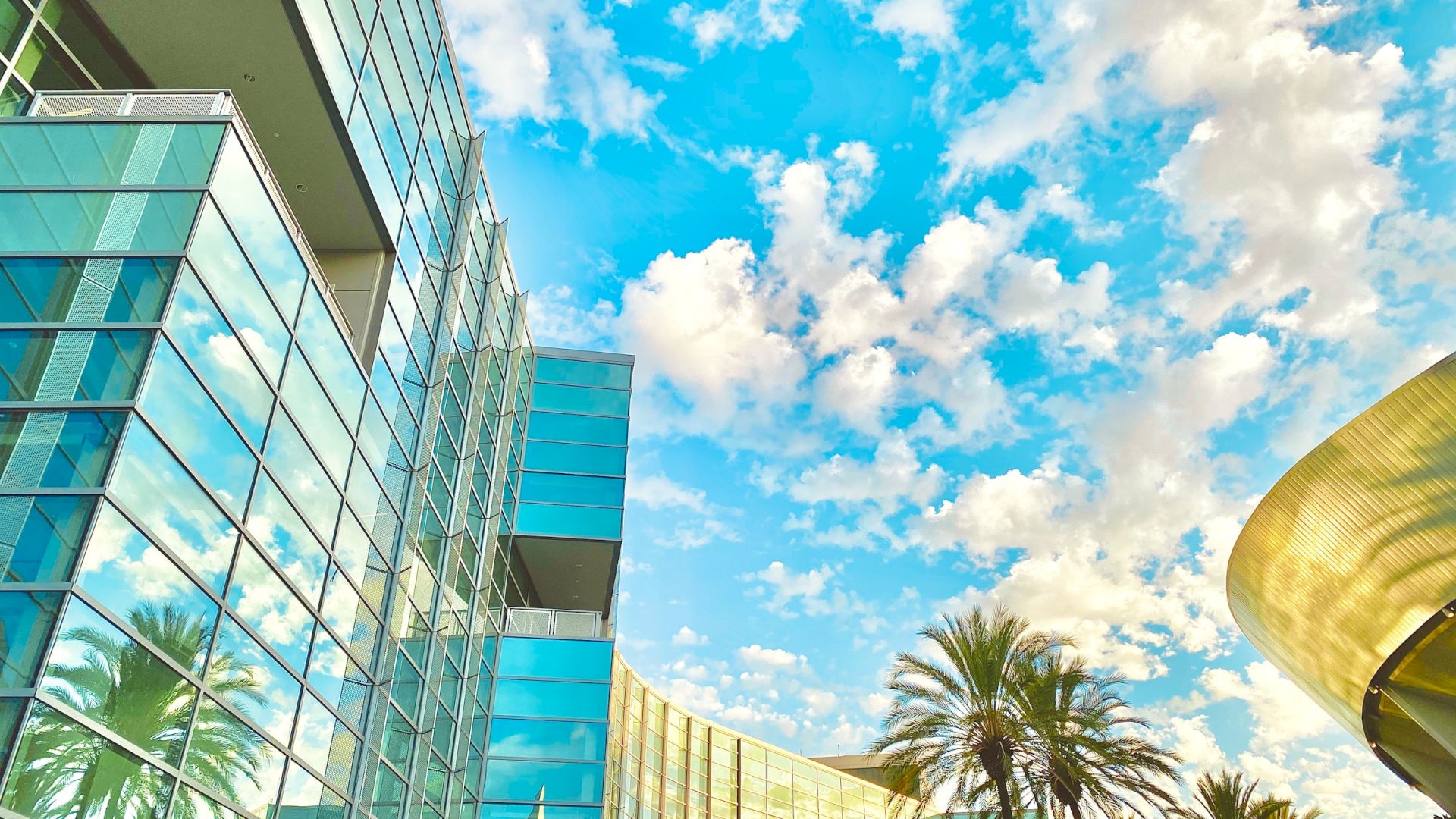The image shows a modern glass building with palm trees in front, a blue sky with scattered clouds, and a curved golden structure to the right.