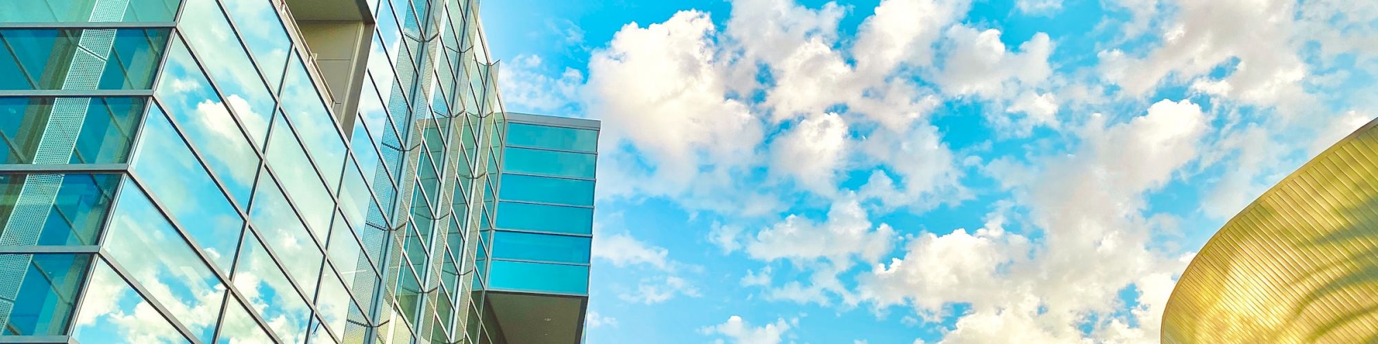 The image shows a modern glass building with palm trees in front, a blue sky with scattered clouds, and a curved golden structure to the right.