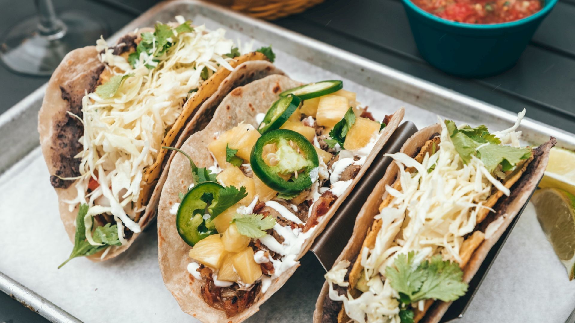 The image shows three tacos on a tray, topped with cheese, jalape&ntilde;os, and other ingredients, along with chips and salsa, and two other tacos in the background.