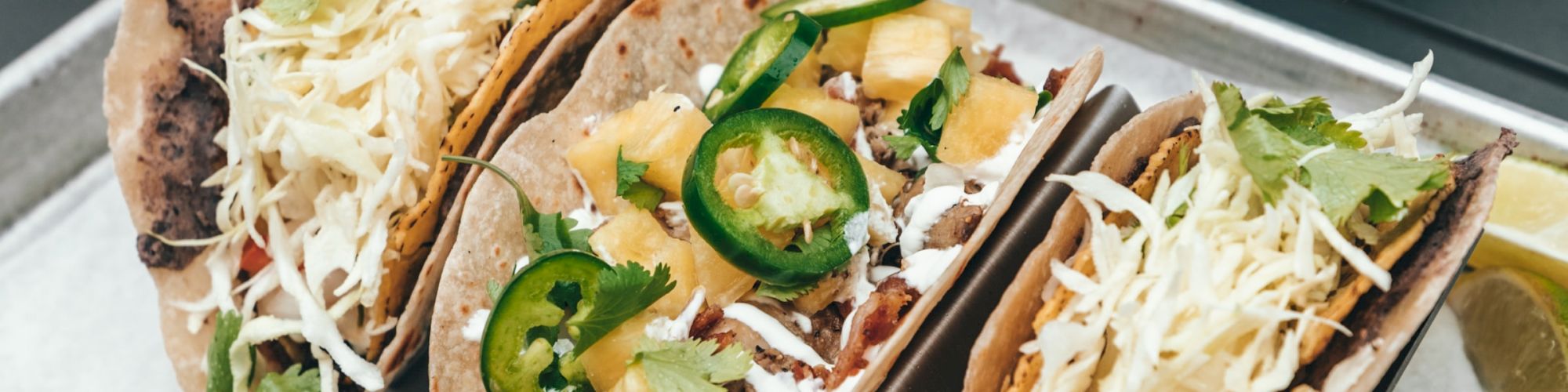 The image shows three tacos on a tray, topped with cheese, jalapeños, and other ingredients, along with chips and salsa, and two other tacos in the background.