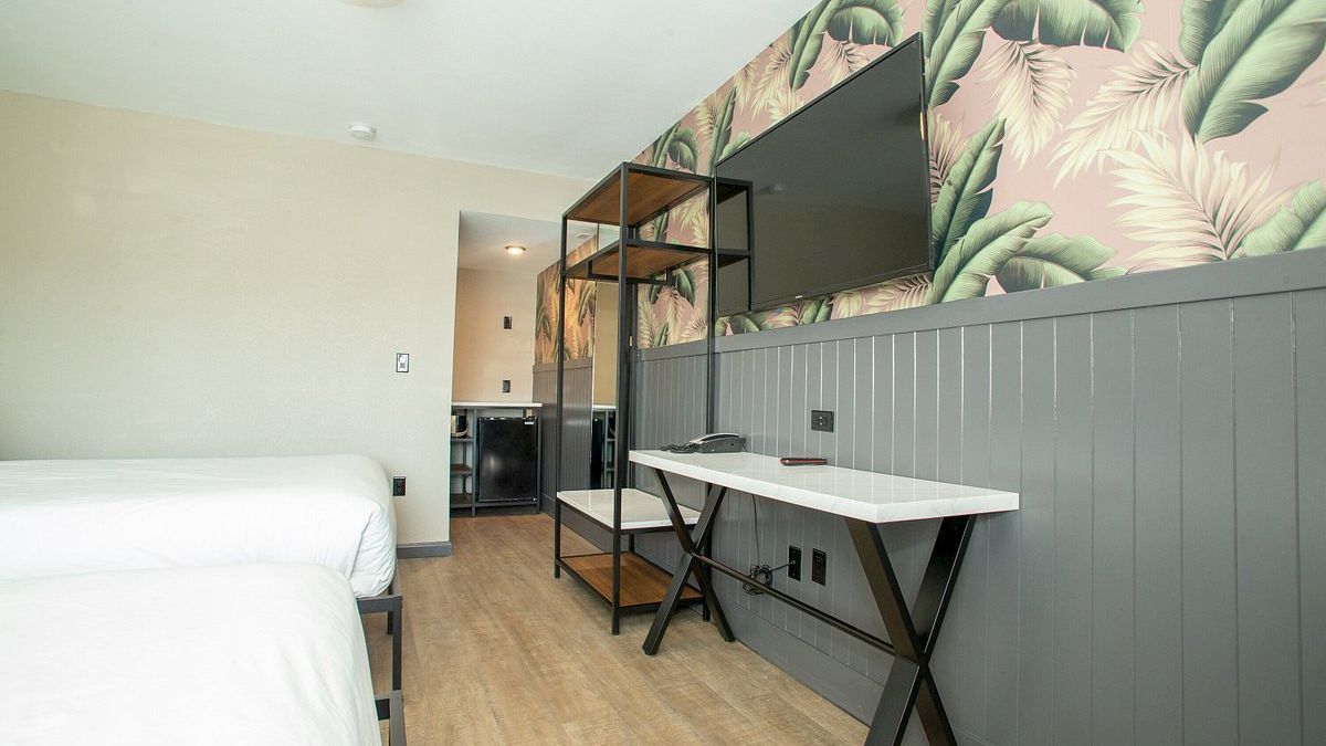 A modern room features two beds, a desk with a chair, a wall with tropical leaf wallpaper, and a shelf. There&rsquo;s a doorway leading to another area.