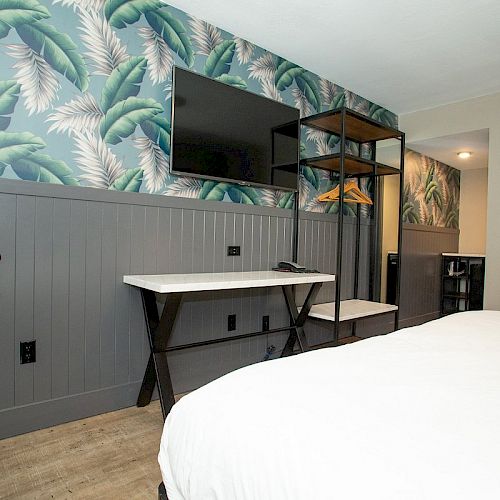 A modern bedroom features a desk, wall-mounted TV, an open wardrobe, and tropical-themed wallpaper with banana leaves, and white bedding.