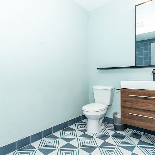 A modern bathroom with white toilet, wooden vanity with sink, large mirror, and blue geometric patterned floor tiles.