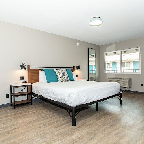 A minimalist bedroom features a bed with blue and white pillows, wall-mounted lamps, a window, a wall-mounted television, a small table, and wooden flooring.