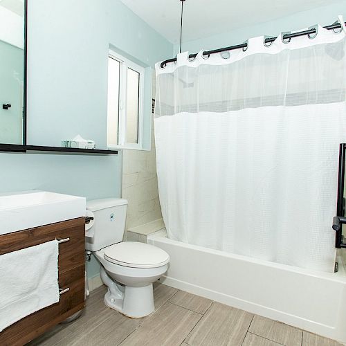 A clean, modern bathroom with a vanity, toilet, bathtub, and shower curtain. It has a large mirror, light-colored walls, and a window above the tub.