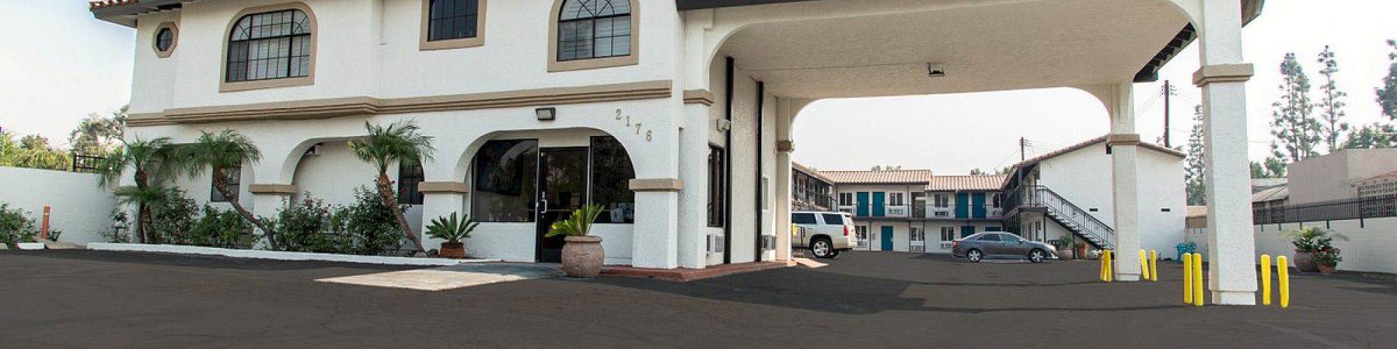 The image shows a building resembling a hotel or motel with an arched entrance, parking lot, and staircase leading to an upper floor under a cloudy sky.