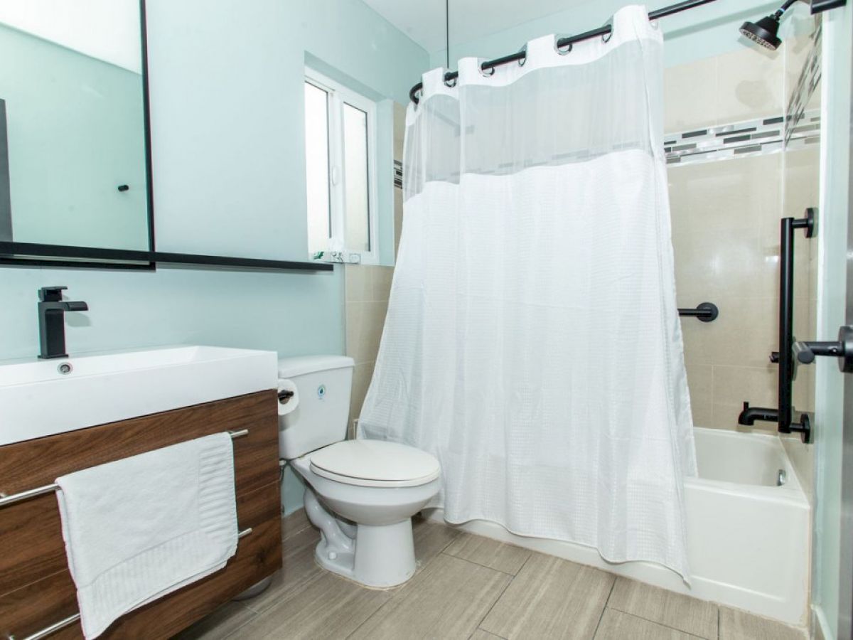 This image shows a modern bathroom with a sink, toilet, and a bathtub with a white shower curtain, along with a mirror above the sink.