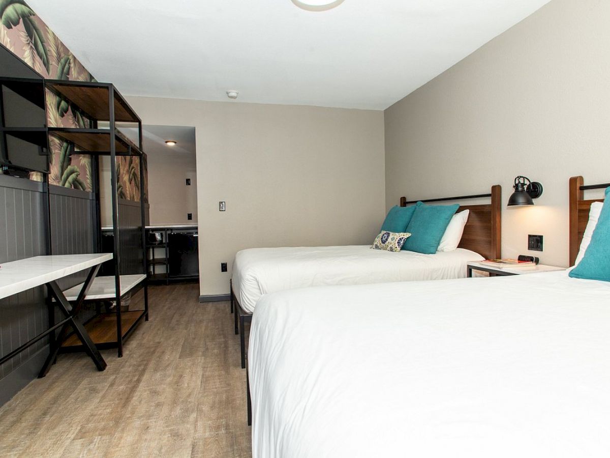 The image shows a modern hotel room with two beds, turquoise pillows, a desk, and an open shelving unit against the wall.