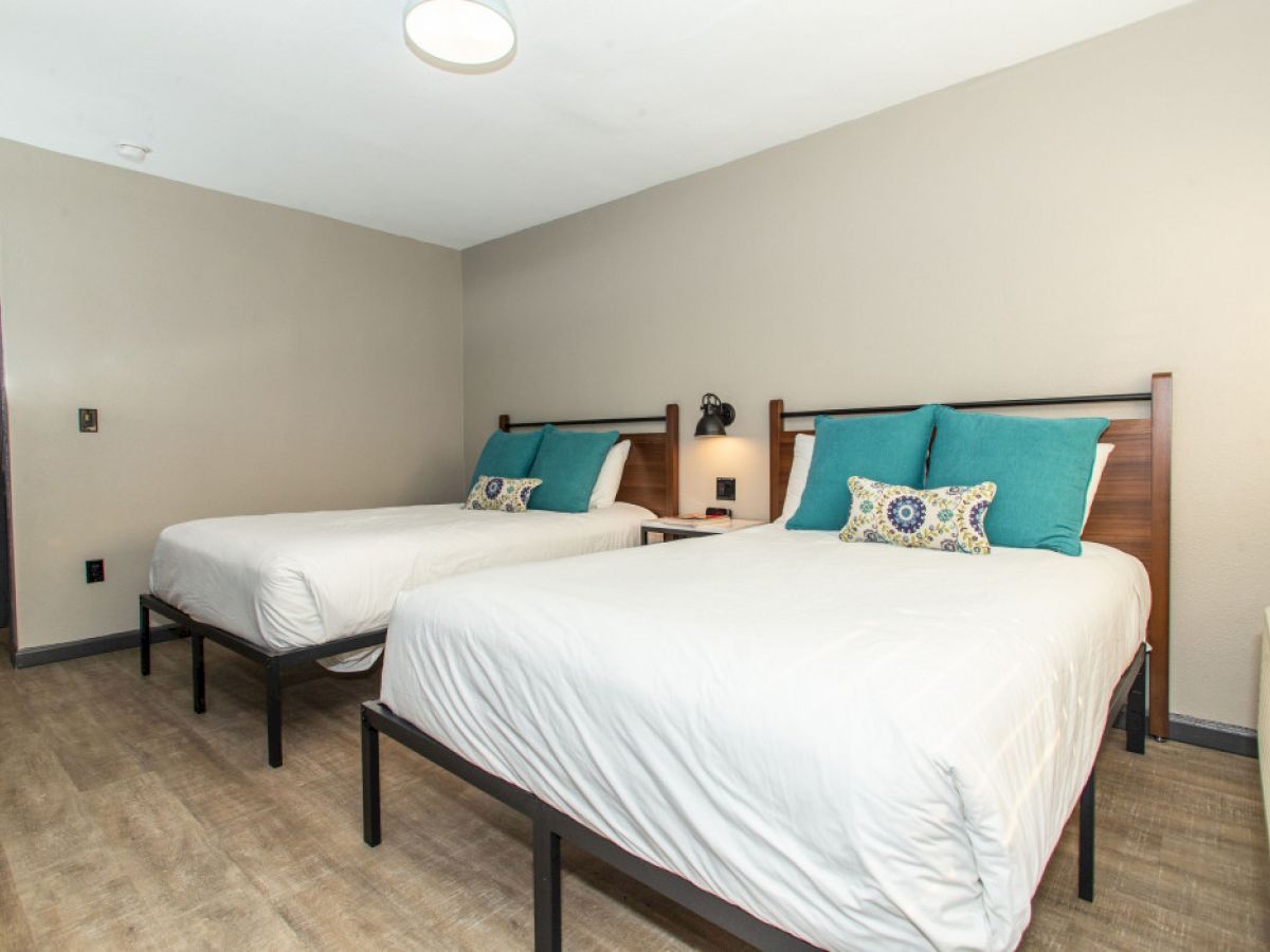 This image shows a hotel room with two double beds, wooden headboards, teal pillows, and a nightstand in between. The walls are beige.