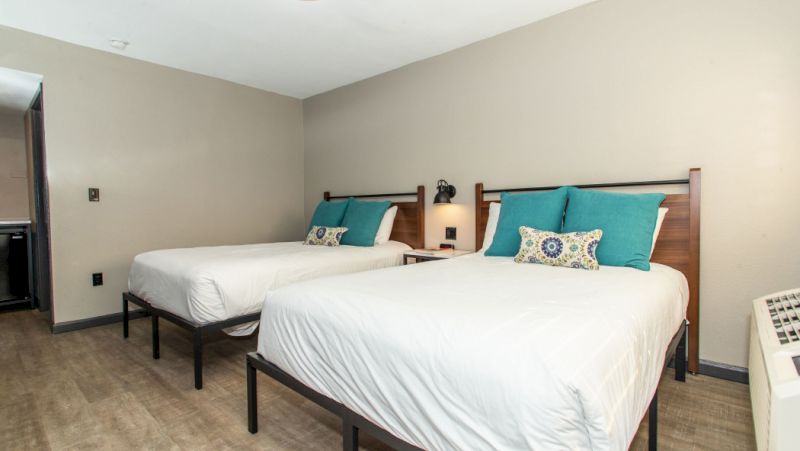 A hotel room with two beds, a nightstand, and teal accent pillows.