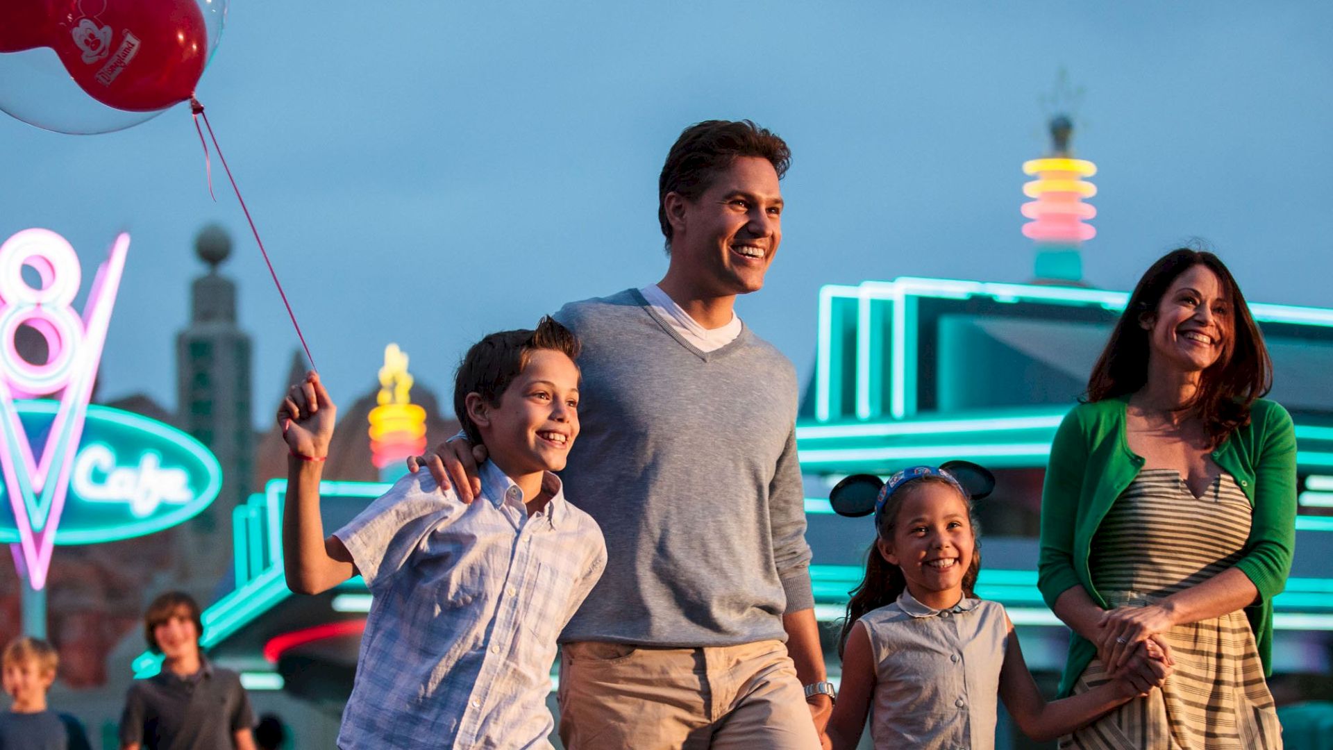 A family of four, holding hands, walks near a brightly lit diner named "Flo's V8 Cafe" at dusk, with the child holding a red balloon.