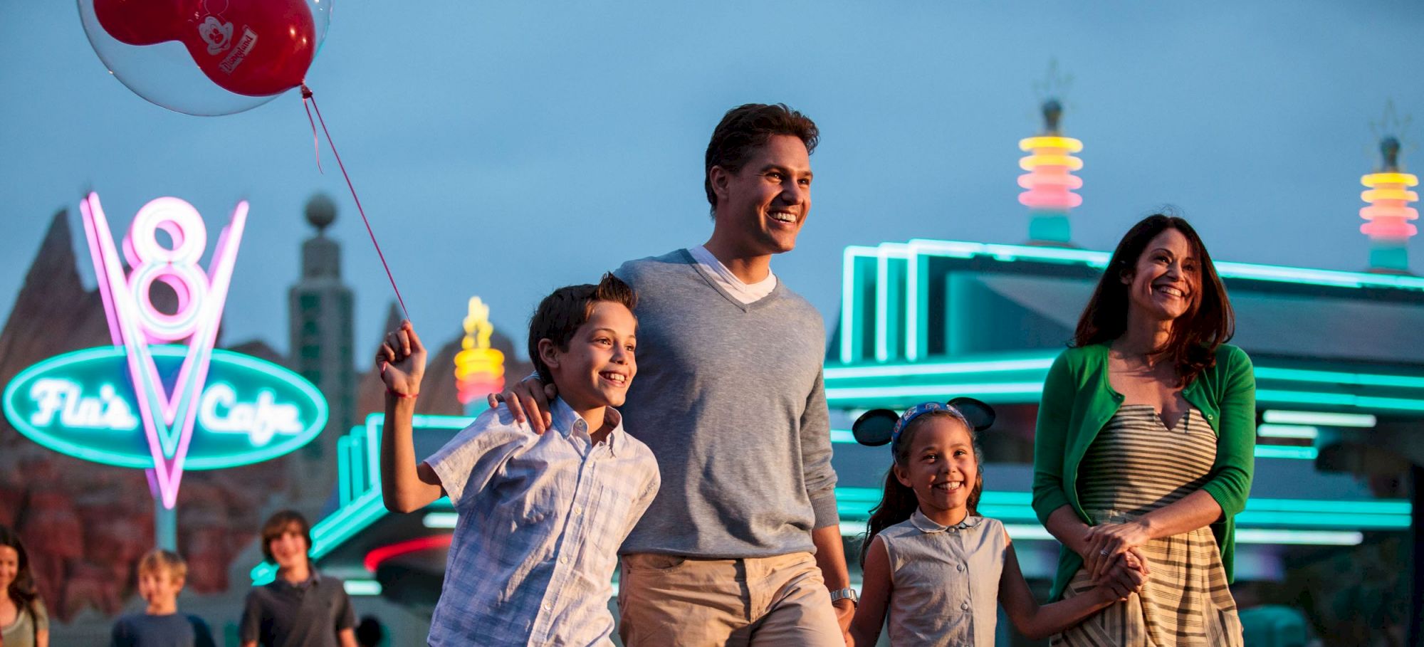 A family of four, holding hands, walks near a brightly lit diner named "Flo's V8 Cafe" at dusk, with the child holding a red balloon.