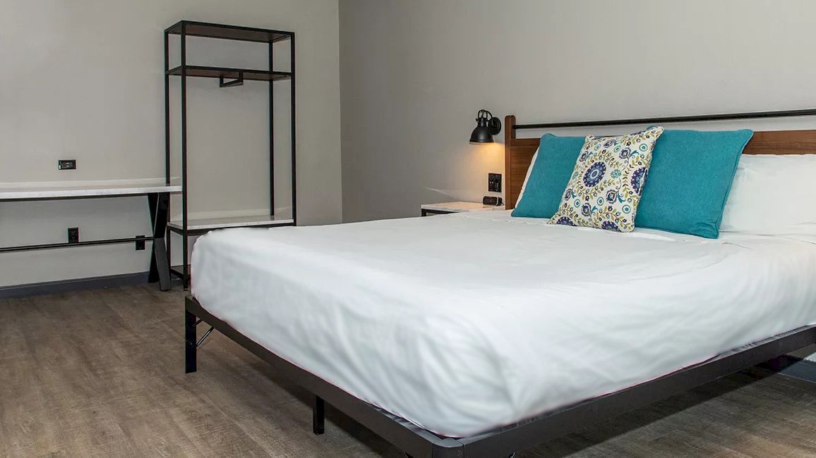 The image shows a modern bedroom with a neatly made bed, blue and patterned pillows, a desk, shelves, and wall-mounted lamps.