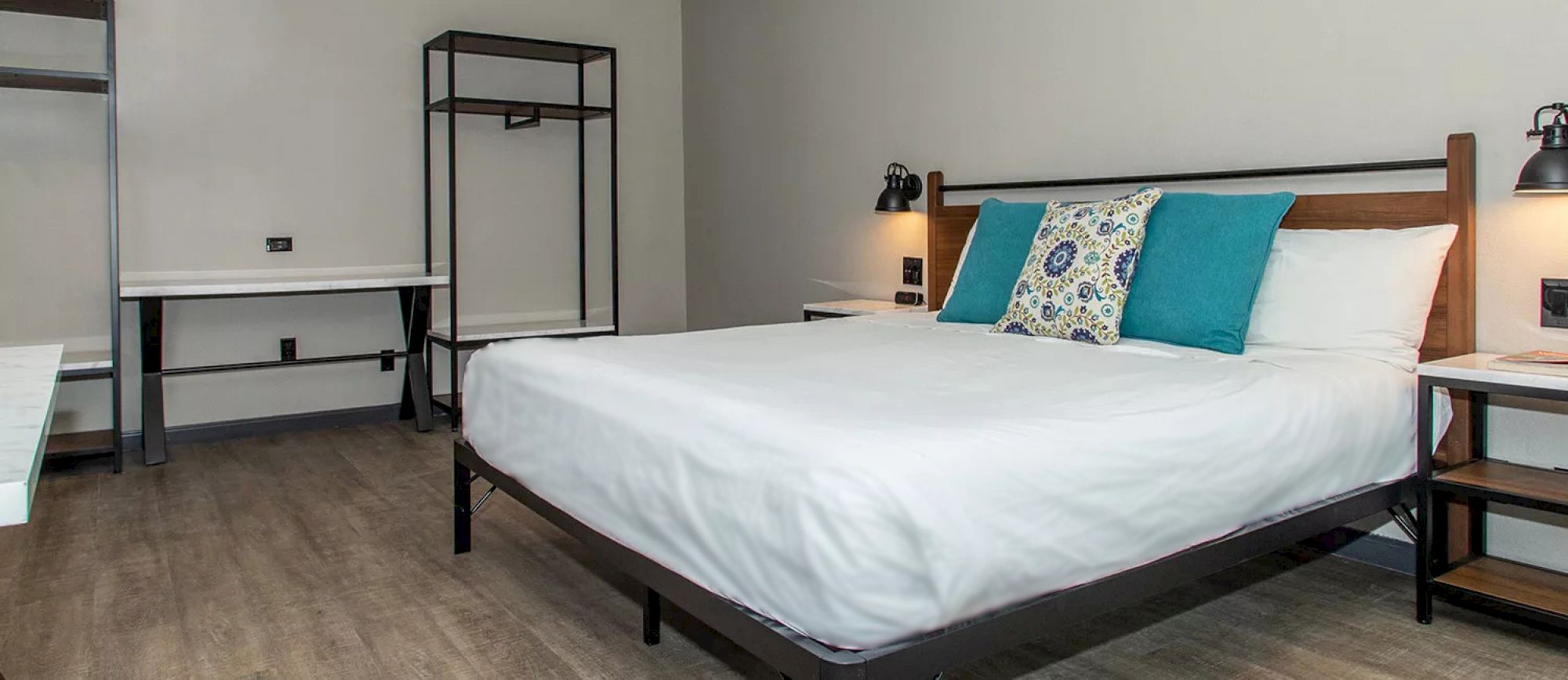 The image shows a modern bedroom with a neatly made bed, blue and patterned pillows, a desk, shelves, and wall-mounted lamps.