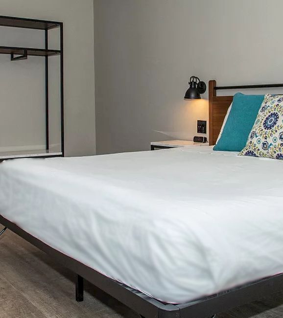A neatly made bed with white sheets and two pillows, one plain teal and one patterned, against a wooden headboard with a black reading light.