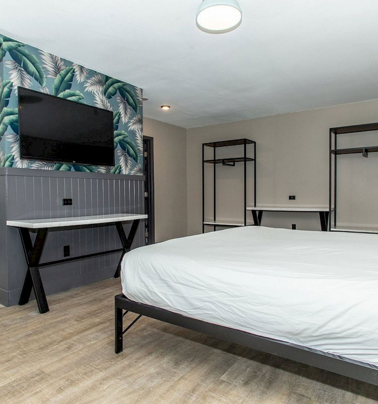 A modern bedroom with a large bed, a TV mounted on a tropical-themed wall, a desk, and a bunk bed in the corner, featuring minimalist decor.