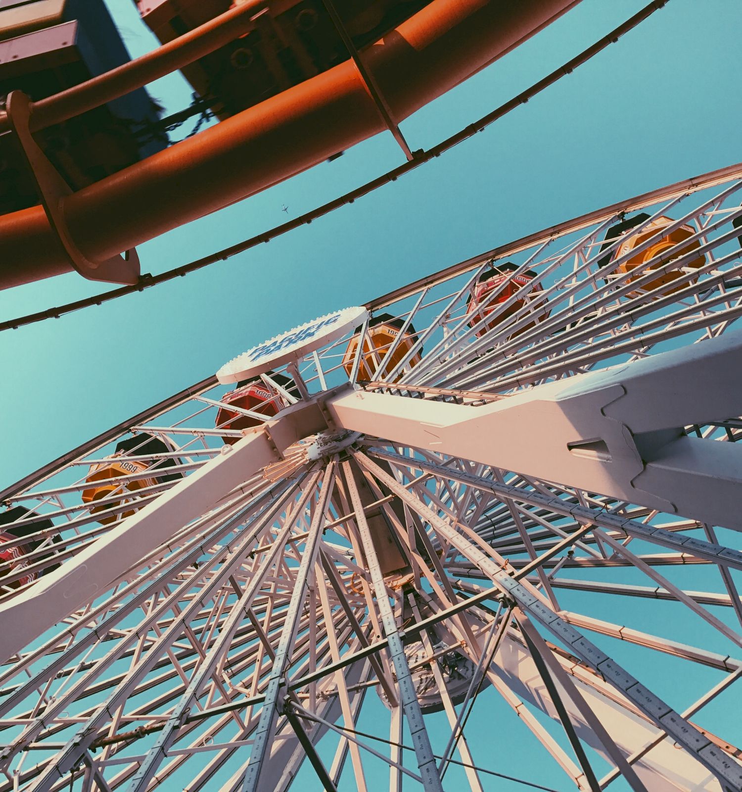 The image shows a view from below of a Ferris wheel with colorful cabins and part of a roller coaster track under a clear sky, creating a fun fair atmosphere.