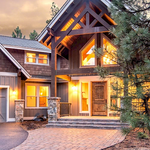 This image shows a large, illuminated house in a wooded area, featuring a three-car garage, wooden and stone elements, and warm, welcoming lights.