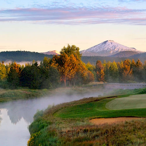 A picturesque landscape with a river, a golf course, trees, and a snow-capped mountain in the background, under a partially cloudy sky.