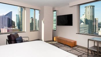 A modern hotel room with large windows, a city skyline view, a bed, a chair, a flat-screen TV, and a wooden bench in front of the TV.