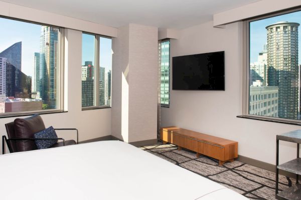 A hotel room with a city view, featuring a bed, chair, TV, and window.