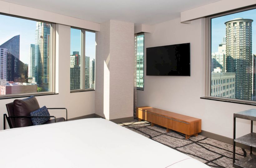 A modern hotel room with large windows, a city skyline view, a bed, a chair, a flat-screen TV, and a wooden bench in front of the TV.