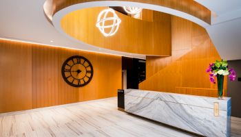 A modern lobby with wooden walls, a marble reception desk, a large wall clock, and a spiral staircase with hanging spherical lights.