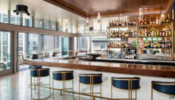 A modern bar with a long counter, stylish chairs, and a well-stocked display of bottles. Large windows provide a view of the city.