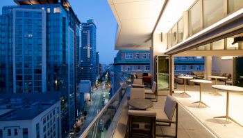 The image shows a city view from a modern balcony with seating areas and tables at twilight, overlooking tall buildings and a street below.